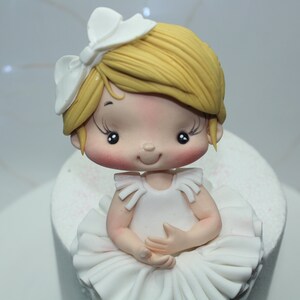 May include: A white fondant ballerina cake topper with a white bow, blonde hair, and a white tutu.