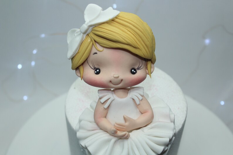 May include: A white fondant ballerina cake topper with a white bow, blonde hair, and a white tutu.