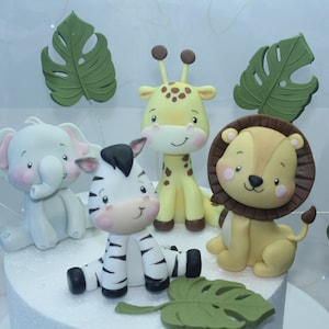 Könnte beinhalten: Ein Set aus fünf Tortendekorationen mit einem Zebra, einer Giraffe, einem Elefanten, einem Löwen und grünen Monstera-Blättern. Die Tiere sind alle cartoonartig und haben freundliche Gesichter. Die Tortendekorationen sind aus Fondant und perfekt für eine Dschungelparty.