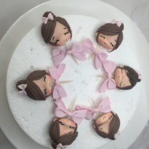 Puede incluir: Seis cabezas de fondant marr&oacute;n de ni&ntilde;a con lazos rosas en la parte superior de los palillos de dientes. Las cabezas est&aacute;n dispuestas en c&iacute;rculo sobre una tabla de pastel blanca.