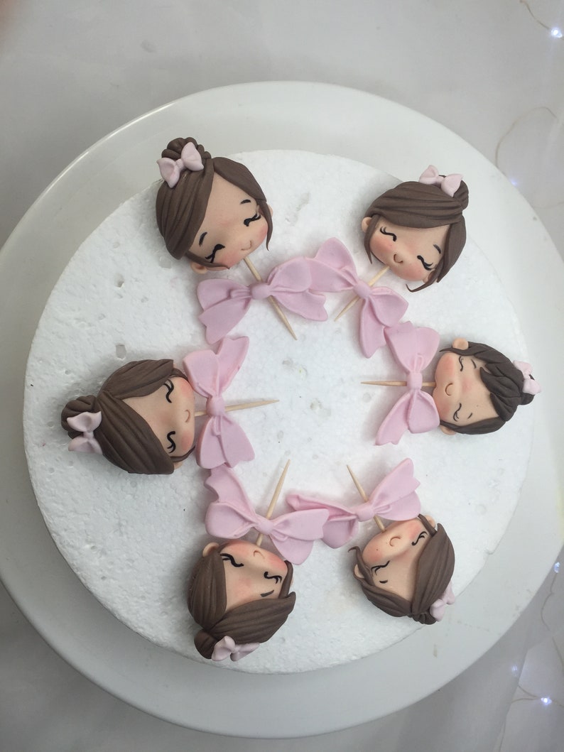 Puede incluir: Seis cabezas de fondant marr&oacute;n de ni&ntilde;a con lazos rosas en la parte superior de los palillos de dientes. Las cabezas est&aacute;n dispuestas en c&iacute;rculo sobre una tabla de pastel blanca.