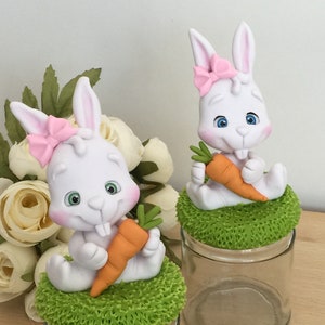 Peut inclure: Deux figurines de lapin en céramique blanche tenant des carottes, chacune assise sur une base verte en forme d'herbe, au-dessus de bocaux en verre transparent. Les lapins ont des nœuds roses et des yeux bleus.