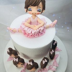 Puede incluir: Un pastel blanco de dos niveles decorado con una mu&ntilde;eca de bailarina de fondant. La bailarina lleva un tut&uacute; rosa y tiene el pelo casta&ntilde;o. El pastel tambi&eacute;n est&aacute; decorado con cabezas de bailarina de fondant m&aacute;s peque&ntilde;as alrededor del nivel inferior.