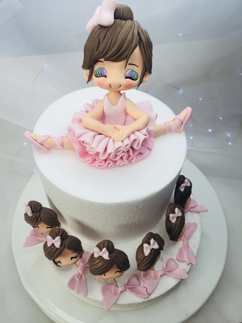 Puede incluir: Un pastel blanco de dos niveles decorado con una mu&ntilde;eca de bailarina de fondant. La bailarina lleva un tut&uacute; rosa y tiene el pelo casta&ntilde;o. El pastel tambi&eacute;n est&aacute; decorado con cabezas de bailarina de fondant m&aacute;s peque&ntilde;as alrededor del nivel inferior.