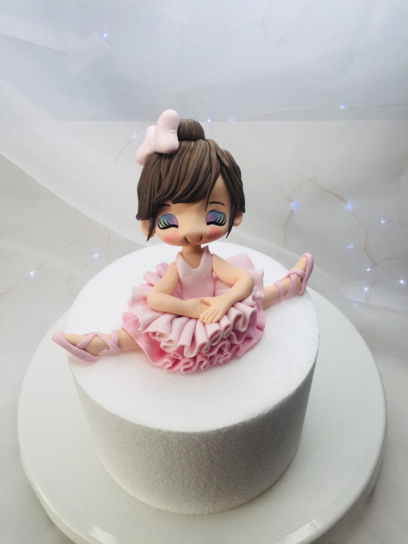 Puede incluir: Un pastel blanco con una figurita de bailarina de fondant en la parte superior. La bailarina lleva un tut&uacute; rosa y tiene el pelo casta&ntilde;o con un lazo rosa.