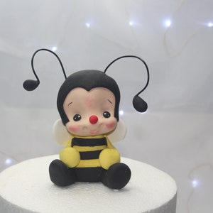 Könnte beinhalten: Eine kleine, schwarz-gelbe Bienenfigur mit freundlichem Gesicht. Die Biene sitzt auf einem weißen Kuchenständer.