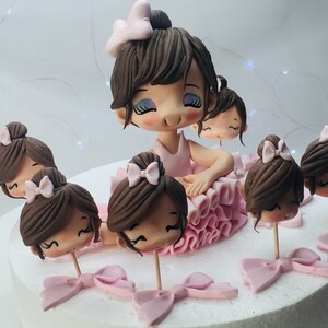 Puede incluir: Un pastel blanco decorado con figuras de bailarinas de fondant. Las figuras llevan todas tut&uacute;s rosas y tienen el pelo casta&ntilde;o. El pastel est&aacute; decorado con lazos rosas y blancos.