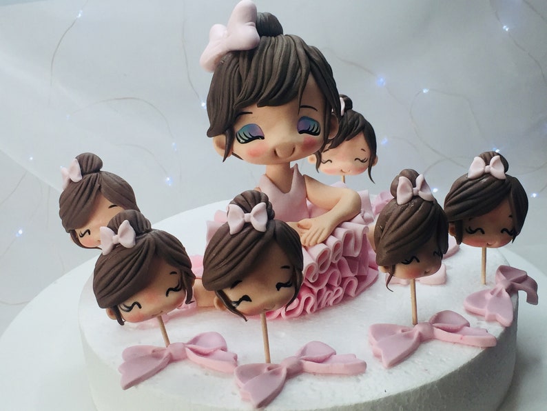 Puede incluir: Un pastel blanco decorado con figuras de bailarinas de fondant. Las figuras llevan todas tut&uacute;s rosas y tienen el pelo casta&ntilde;o. El pastel est&aacute; decorado con lazos rosas y blancos.