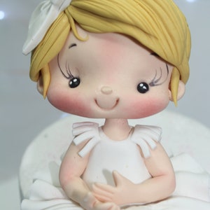 May include: A white fondant ballerina cake topper with a white bow, blonde hair, and a white dress. The ballerina has a smiling face with rosy cheeks and closed eyes.