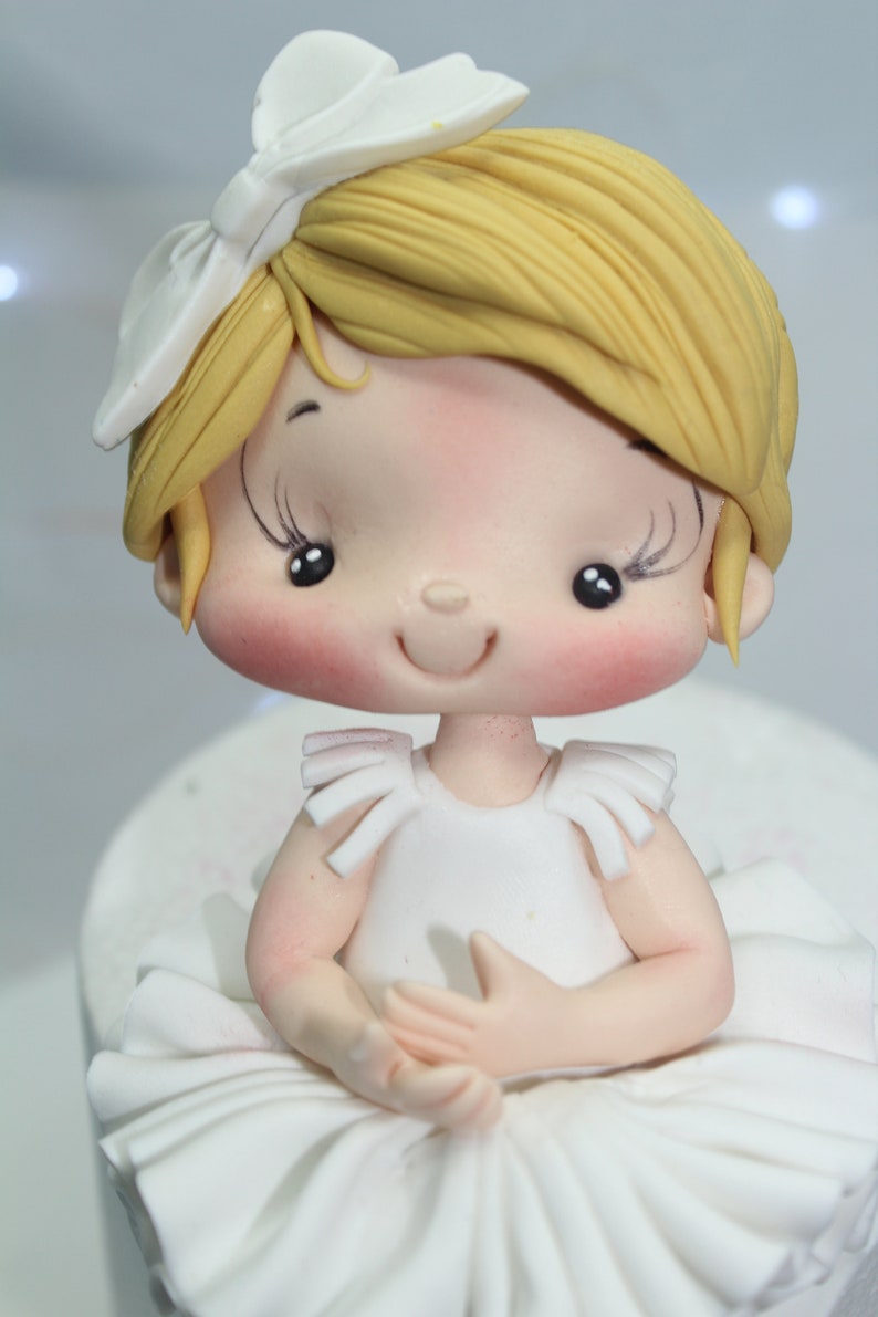 May include: A white fondant ballerina cake topper with a white bow, blonde hair, and a white dress. The ballerina has a smiling face with rosy cheeks and closed eyes.