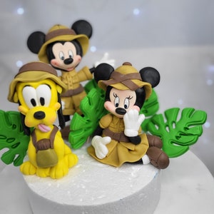 Puede incluir: Un conjunto de adornos para tartas con Mickey Mouse, Minnie Mouse y Pluto vestidos con atuendos de safari. Los personajes están hechos de fondant y están rodeados de hojas verdes.