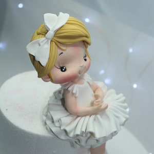 May include: A white fondant ballerina cake topper with a white bow in her hair and a white tutu. She has blonde hair and rosy cheeks.
