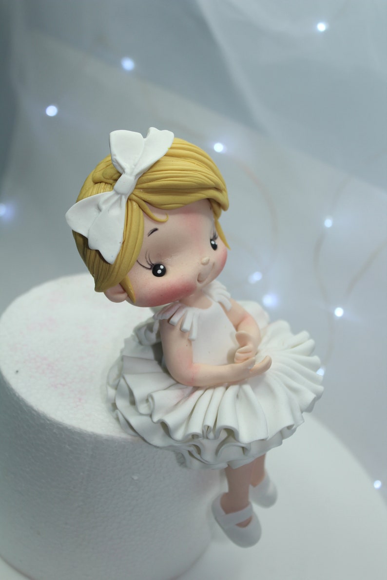 May include: A white fondant ballerina cake topper with a white bow in her hair and a white tutu. She has blonde hair and rosy cheeks.