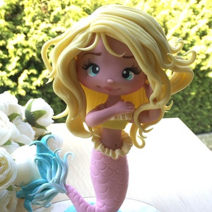 May include: A handcrafted mermaid figurine with a light pink scaled tail and a yellow top. The mermaid has long, flowing blonde hair and blue eyes. The figurine is on a clear base and is a decorative item.