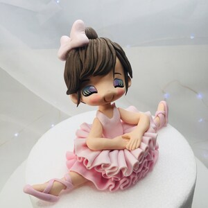 Puede incluir: Un adorno de pastel de bailarina de fondant rosa y marr&oacute;n. La bailarina lleva un tut&uacute; rosa y zapatos de ballet rosas. Tiene el pelo marr&oacute;n y un lazo rosa.
