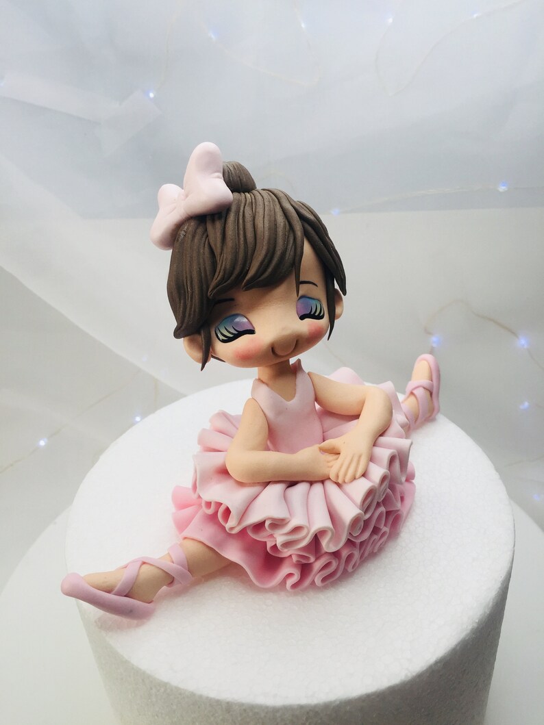 Puede incluir: Un adorno de pastel de bailarina de fondant rosa y marr&oacute;n. La bailarina lleva un tut&uacute; rosa y zapatos de ballet rosas. Tiene el pelo marr&oacute;n y un lazo rosa.