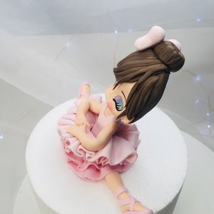 Puede incluir: Un adorno de pastel de bailarina rosa con un mo&ntilde;o marr&oacute;n y un lazo rosa. La bailarina lleva un tut&uacute; rosa y zapatos rosas.