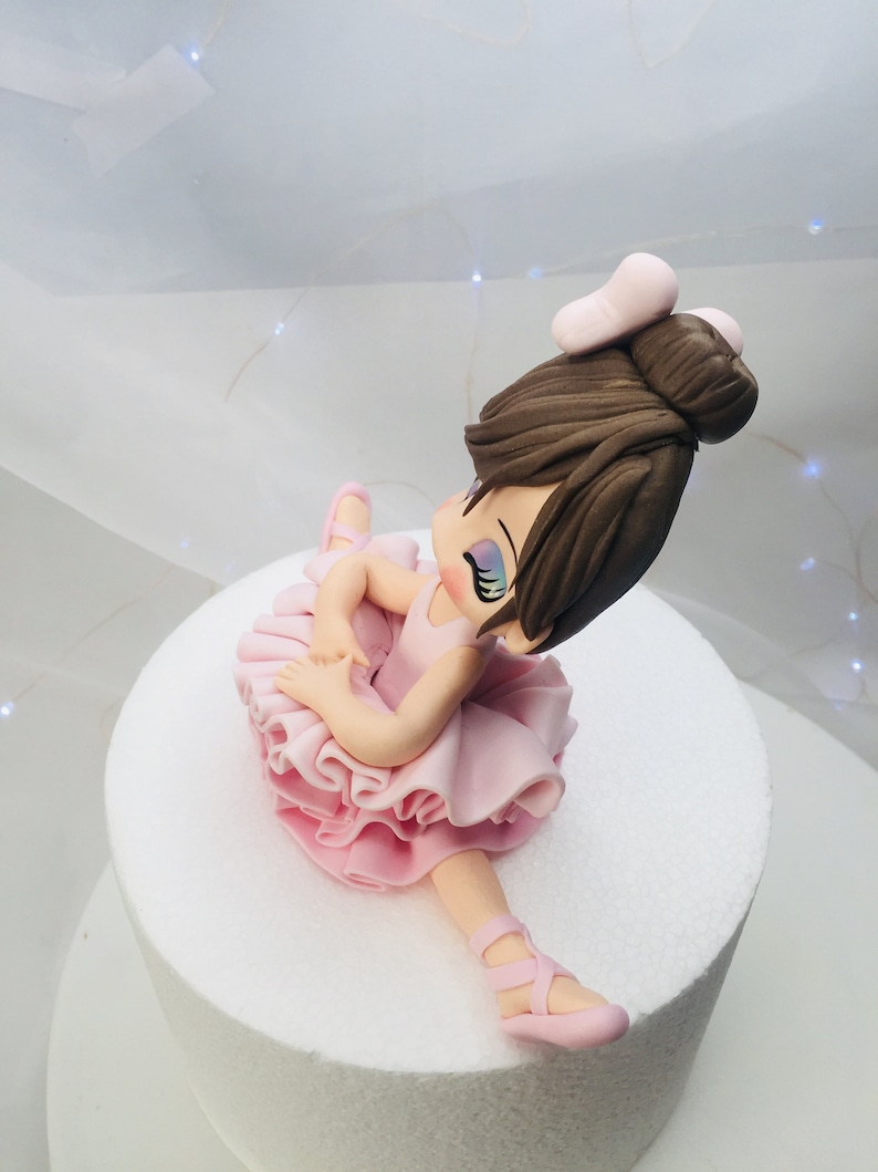 Puede incluir: Un adorno de pastel de bailarina rosa con un mo&ntilde;o marr&oacute;n y un lazo rosa. La bailarina lleva un tut&uacute; rosa y zapatos rosas.
