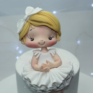 May include: A white fondant ballerina cake topper with a white bow, blonde hair, and a pink face. The ballerina is wearing a white tutu and has her hands clasped in front of her.