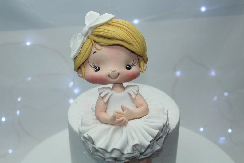May include: A white fondant ballerina cake topper with a white bow, blonde hair, and a pink face. The ballerina is wearing a white tutu and has her hands clasped in front of her.