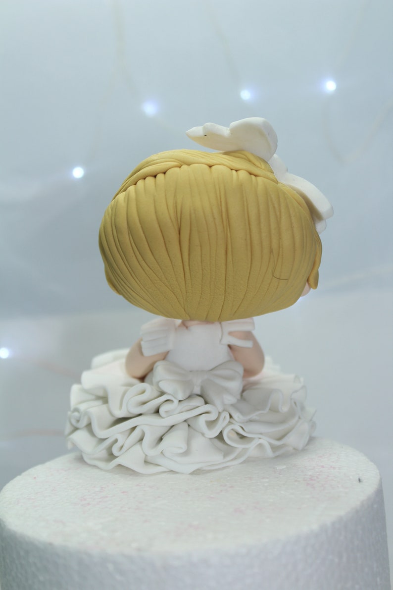 May include: A white fondant cake topper featuring a small girl with blonde hair wearing a white dress with a ruffled skirt. The girl is sitting on the cake and has her back to the viewer.