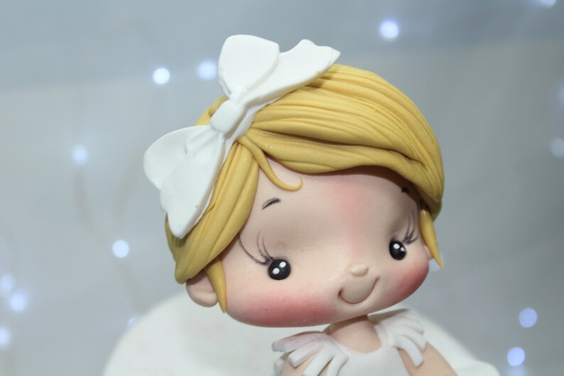 May include: A close-up of a fondant figurine of a girl with blonde hair, wearing a white bow and a white dress. The figurine has a smiling face with rosy cheeks and large, dark eyes.