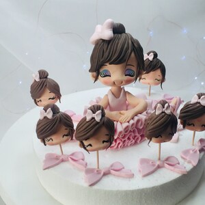 Puede incluir: Un pastel de fondant rosa y marr&oacute;n decorado con una figura de bailarina y varias cabezas de bailarina m&aacute;s peque&ntilde;as. La figura de bailarina lleva un tut&uacute; rosa y tiene el pelo marr&oacute;n. Las cabezas de bailarina m&aacute;s peque&ntilde;as tambi&eacute;n llevan tut&uacute;s rosas y tienen el pelo marr&oacute;n.