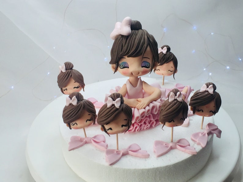 Puede incluir: Un pastel de fondant rosa y marr&oacute;n decorado con una figura de bailarina y varias cabezas de bailarina m&aacute;s peque&ntilde;as. La figura de bailarina lleva un tut&uacute; rosa y tiene el pelo marr&oacute;n. Las cabezas de bailarina m&aacute;s peque&ntilde;as tambi&eacute;n llevan tut&uacute;s rosas y tienen el pelo marr&oacute;n.