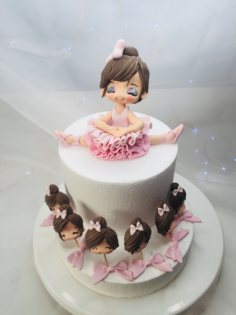 Puede incluir: Un pastel blanco decorado con una mu&ntilde;eca de bailarina de fondant y mu&ntilde;ecas de bailarina de fondant m&aacute;s peque&ntilde;as alrededor de la base del pastel.