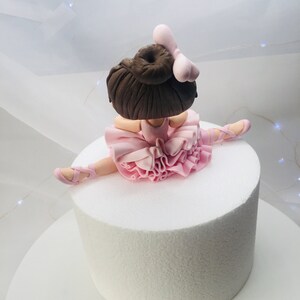 Puede incluir: Un adorno de pastel de bailarina de fondant rosa y marr&oacute;n. La bailarina lleva un tut&uacute; rosa y tiene las piernas en una abertura. Tiene el pelo marr&oacute;n y un lazo rosa.