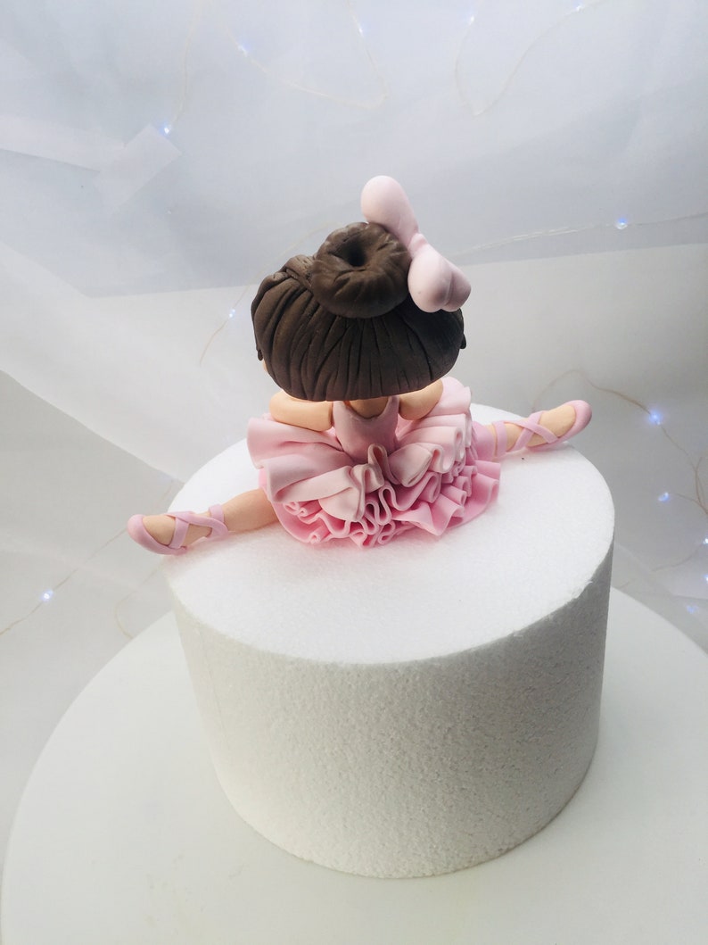 Puede incluir: Un adorno de pastel de bailarina de fondant rosa y marr&oacute;n. La bailarina lleva un tut&uacute; rosa y tiene las piernas en una abertura. Tiene el pelo marr&oacute;n y un lazo rosa.