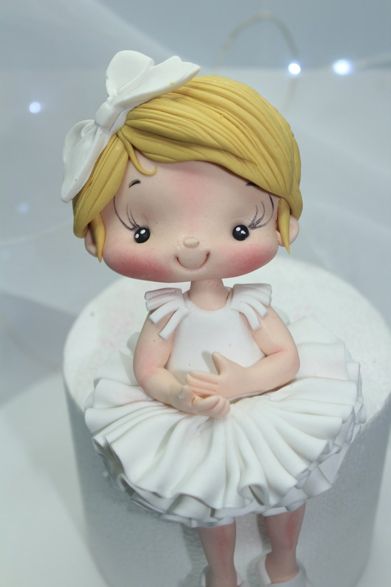 May include: A white fondant ballerina figurine with blonde hair, a white bow, and a white tutu. The figurine is sitting on a white cake.