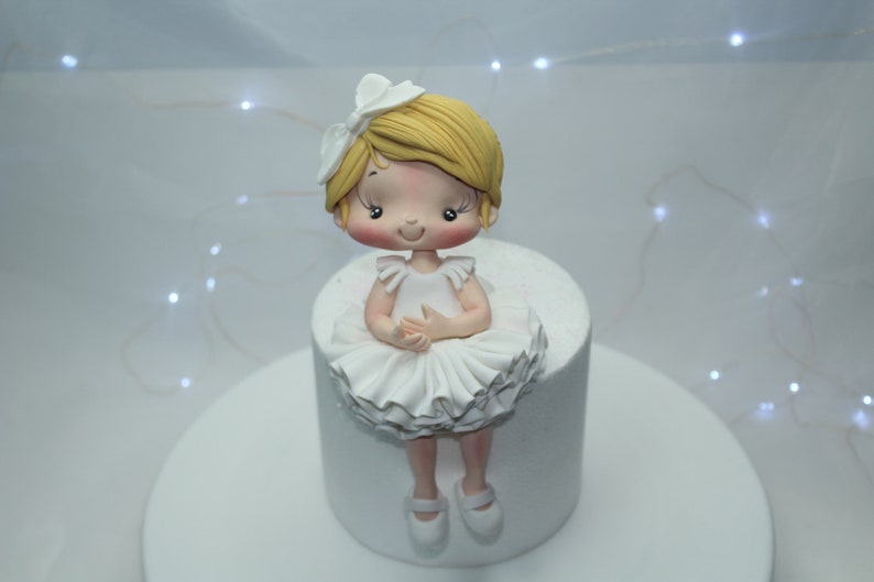 May include: A white cake with a fondant ballerina figurine on top. The ballerina has blonde hair, a white dress, and pink shoes.