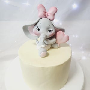 Könnte beinhalten: Ein weißer Kuchen mit einer grauen Fondant-Elefantenfigur darauf. Der Elefant trägt eine rosa Schleife und hält ein rosa Herz.