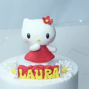 Pode incluir: Topper de bolo Hello Kitty branco com um vestido vermelho e uma flor amarela na cabeça. O bolo é decorado com margaritas brancas e o nome "LAURA" em letras amarelas.