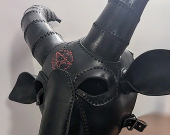 May include: A black leather goat mask with stitched detailing and curved horns. The mask features a red embroidered design on the forehead, eye openings, and ear shapes. A leather strap and fringe complete the design.