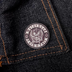 Puede incluir: Un parche redondo de Motorhead gris oscuro con un diseño de calavera y huesos cruzados, rodeado por las palabras "Motorhead" y "Born to Roll". El parche está sobre un fondo de mezclilla oscura con costuras naranjas.