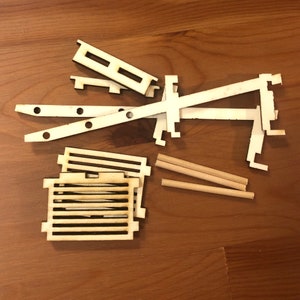 K&ouml;nnte beinhalten: Ein Satz aus Holzbausteinen zum Bau eines Modells. Das Set enth&auml;lt verschiedene Formen und Gr&ouml;&szlig;en von Holzst&uuml;cken, darunter zwei lange St&uuml;cke, zwei kurze St&uuml;cke und ein quadratisches St&uuml;ck mit einem Gittermuster. Die St&uuml;cke sind alle aus hellem Holz gefertigt.