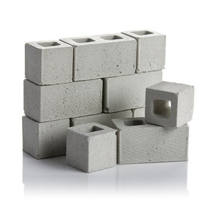 May include: A stack of grey concrete blocks, some arranged in a wall pattern, others stacked on top of each other. The blocks have a rough texture and a hole in the centre.