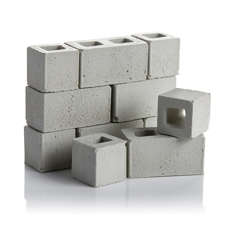 May include: A stack of grey concrete blocks, some arranged in a wall pattern, others stacked on top of each other. The blocks have a rough texture and a hole in the centre.