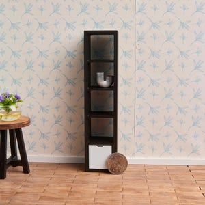 Könnte beinhalten: Ein schwarzes Holzregal mit fünf Regalböden und einer weißen Schublade unten. Das Regal steht an einer Wand mit einem hellblauen Blumenmuster. Eine silberne Metallskulptur steht auf dem obersten Regalboden.