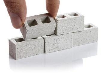 Puede incluir: Una mano coloca un bloque de hormigón gris encima de una pequeña pared hecha de bloques de hormigón grises.