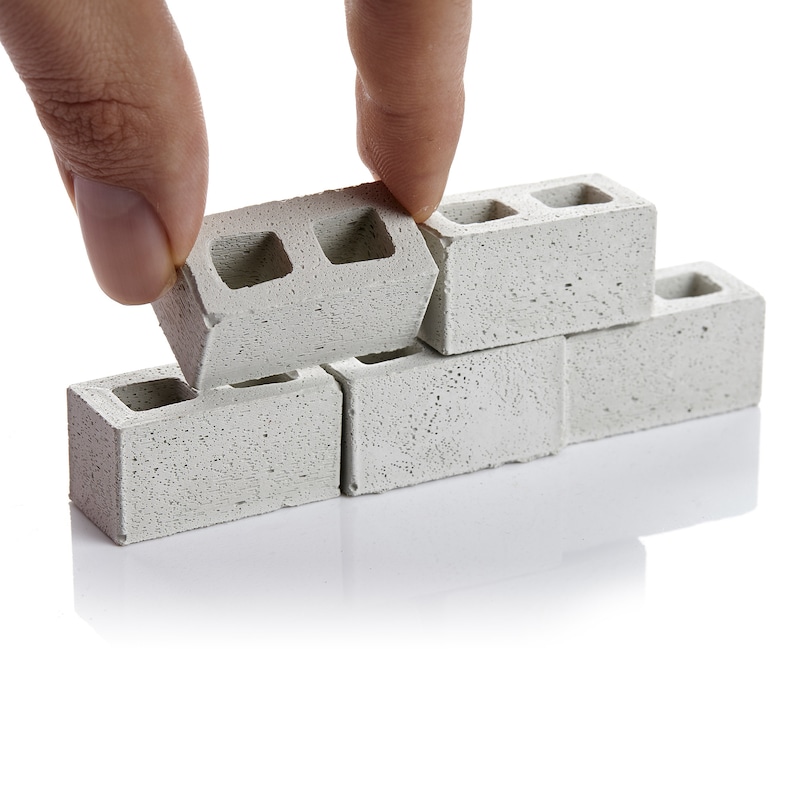 May include: A hand placing a grey concrete block on top of a small wall made of grey concrete blocks.