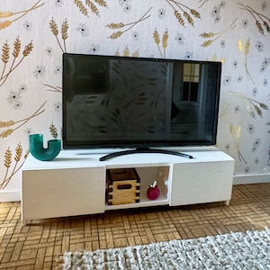 May include: A white TV stand with two drawers and an open shelf. The stand has a flat top surface with a black TV on top. There is a green vase, a wooden box, and a pink vase on the shelf.