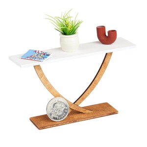 May include: White console table with a wooden base. The table has a plant in a white pot, a red vase, and a book on the top.