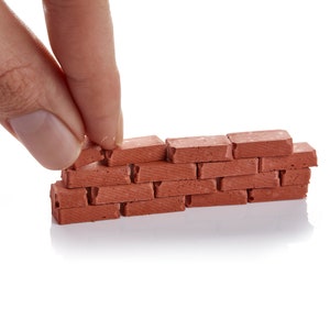 Könnte beinhalten: Eine Hand legt einen kleinen roten Ziegel auf eine Miniatur-Ziegelmauer. Die Mauer besteht aus mehreren roten Ziegeln, die jeweils eine leicht unterschiedliche Textur haben. Die Mauer steht auf einer weißen Oberfläche.