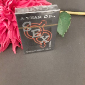 May include: A black card deck with the word "SEX" in orange and silver lettering. The text "A YEAR OF..." is above the word "SEX" and "SEXUAL POSITION CARDS" is below. The deck is next to a pink flower.
