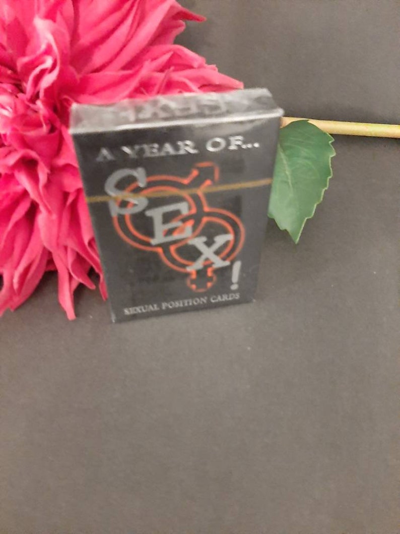 May include: A black card deck with the word "SEX" in orange and silver lettering. The text "A YEAR OF..." is above the word "SEX" and "SEXUAL POSITION CARDS" is below. The deck is next to a pink flower.