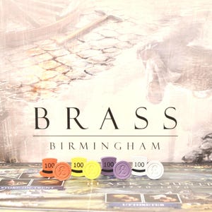 May include: The board game "Brass: Birmingham" is displayed, featuring a backdrop of a waterway and cobblestone street. The game's title is prominently displayed. Colorful game tokens, including orange, yellow, and purple, are arranged in front of the board.
