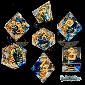 Puede incluir: Conjunto de siete dados azules y dorados con un diseño de dragón. Los dados están hechos de resina transparente con detalles dorados y remolinos azules. Los números en los dados son dorados.
